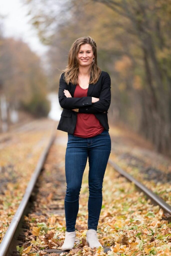Image of Liz as she stands in the middle of train tracks with fall leaves all around her. She is smiling. 