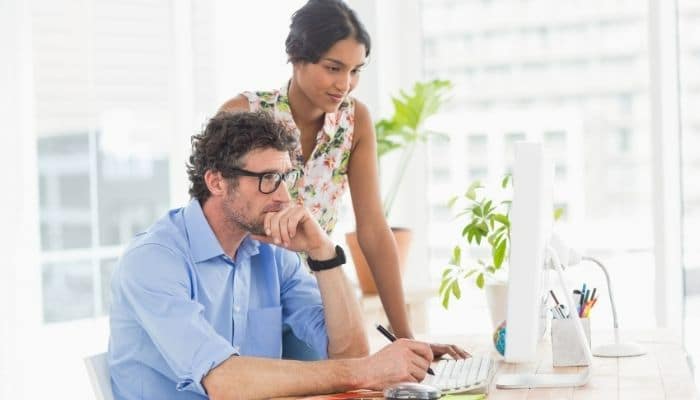 Man sitting at his desk looking at the computer screen with a woman standing behind him looking at his computer screen