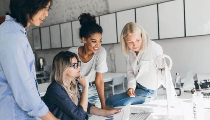 Diverse team of marketing professionals gathered in front of a computer screen. Three women and one man are brainstorming ideas to write email subject lines for event invitations