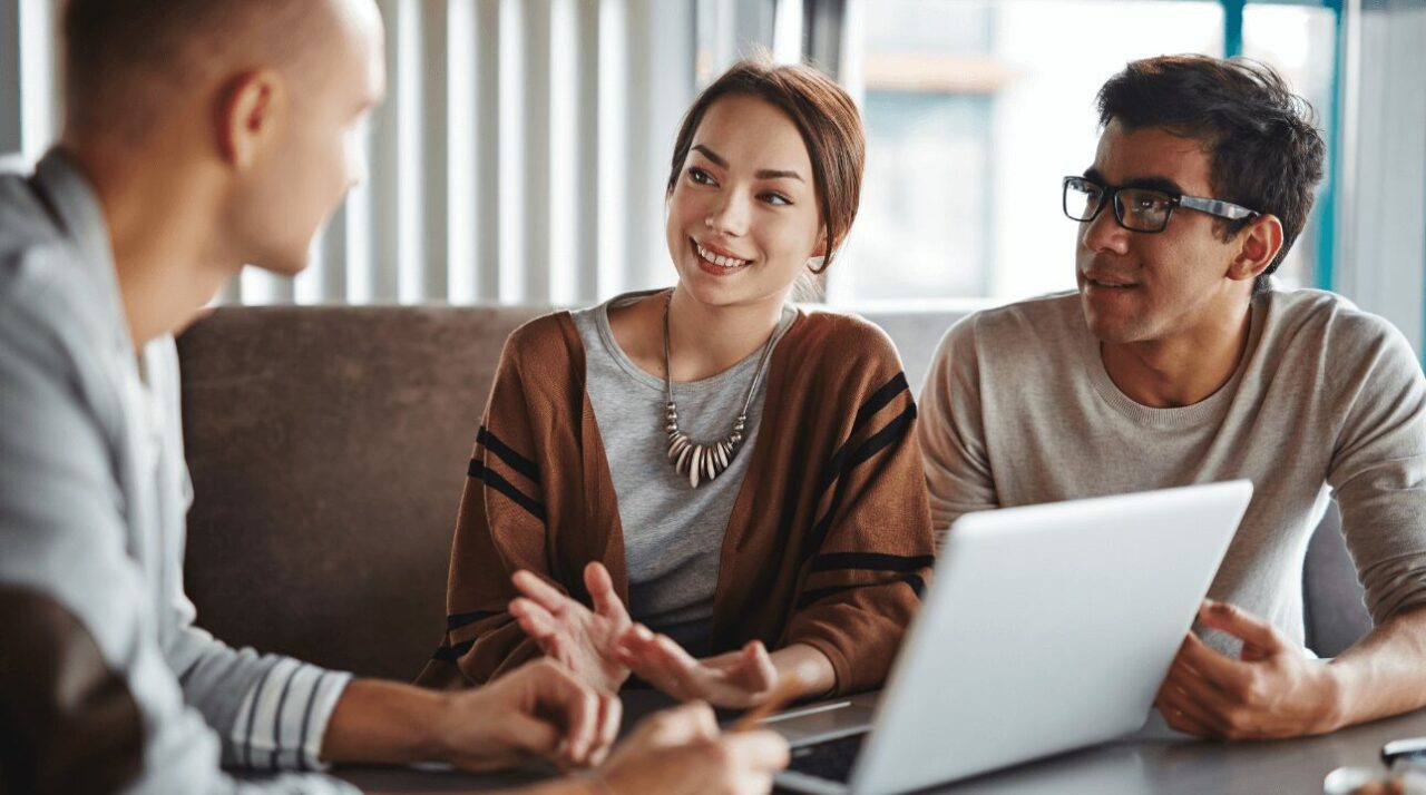 Picture of three coworkers talk in front of a laptop.