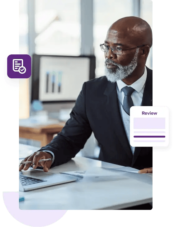 A bald businessman with a grey beard sits at his desk wearing a dark blue suit and reaches out to touch the mousepad on his laptop.