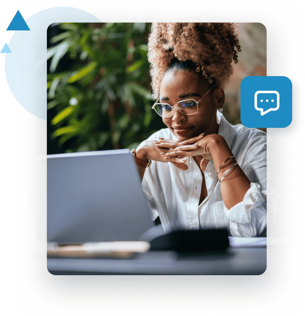 A woman in glasses sits at a desk with her hands crossed under chin and looks at her laptop.