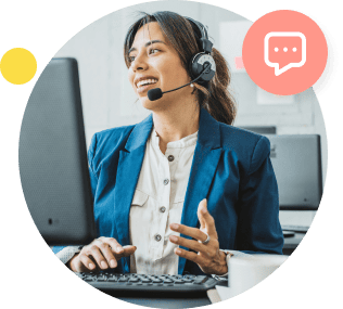 A woman in a blue blazer wears a hands-free headset and talks to a client while seated in front of her PC.