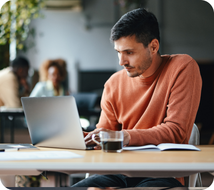 Un hombre con un suéter naranja se sienta en su escritorio con una computadora portátil y una taza de café transparente.