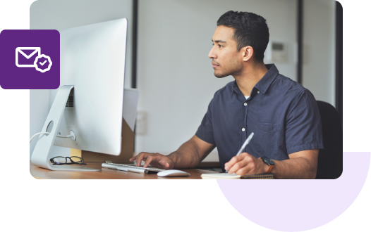 A man in a blue shirt types on his Mac with one hand and writes on a notepad with the other.