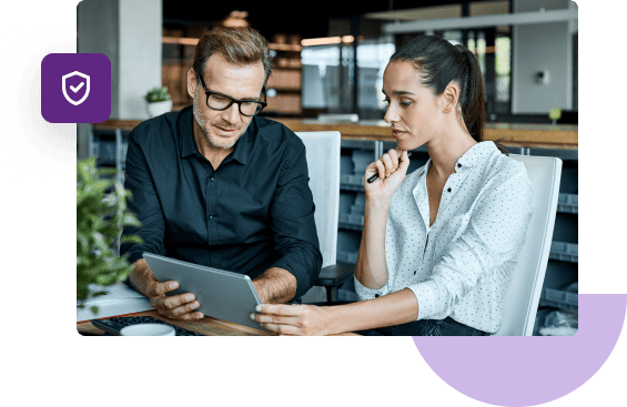 Two business partners sit at a desk and look at a tablet that's held up between them.