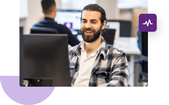 A man in a plaid shirt smiles in front of his computer while another man sits in front of his computer in the background.