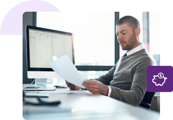 A man in a grey sweater reviews two documents in his hands with a spreadsheet open on his Mac in the background.