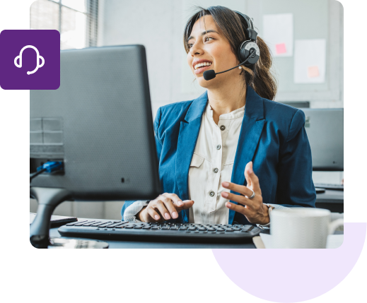 A female customer support agent wearing a hands-free headset is caught mid-speech in front of her keyboard and monitor.