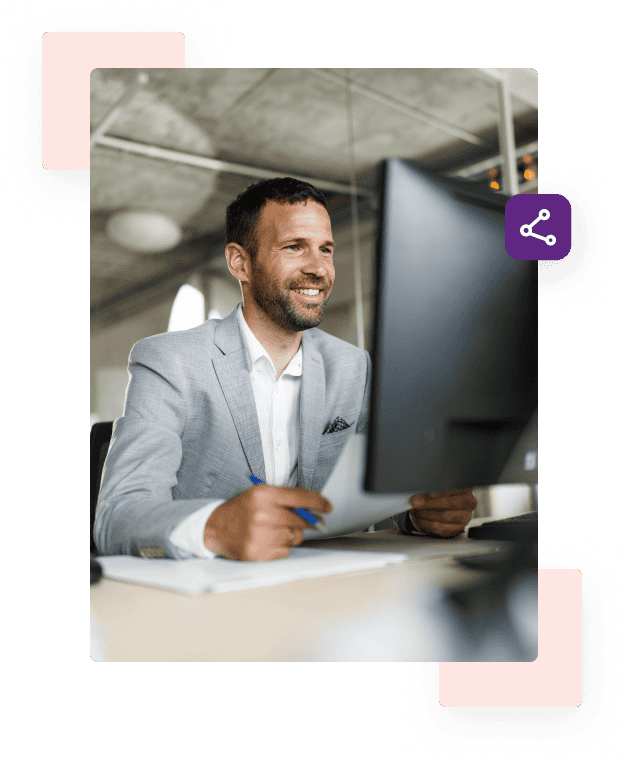 A man in a grey blazer and white shirt smiles in front of his computer monitor and holds a paper and blue pen.