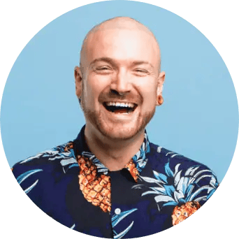 A bald man with gauge earrings wears a blue shirt with pineapples and smiles wide.