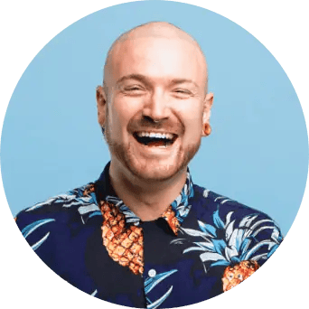A bald man with gauge earrings wears a blue shirt with pineapples and smiles wide.