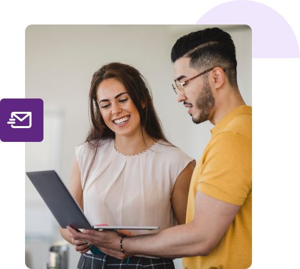 A man holds his laptop and shows his screen to a woman next to him that’s smiling.