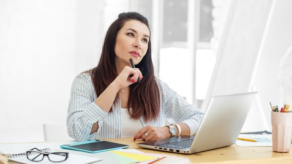 Woman of Asian descent ponders the perfect sales email while sitting in front of laptop computer.