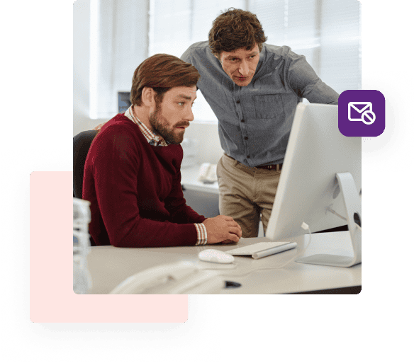 A man in a red sweater seated at his desk looks intently at his computer while another man in a grey shirt stands next to him pointing at the screen.