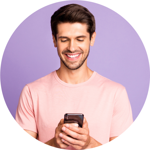 A man in a pink t-shirt smiles as he looks down at his mobile phone.