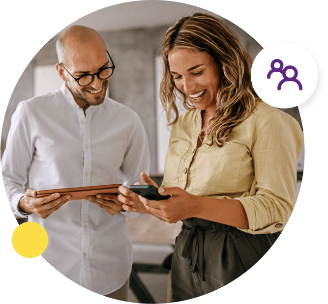 A man holding a tablet smiles as he looks over at a mobile that his female business partner is holding.